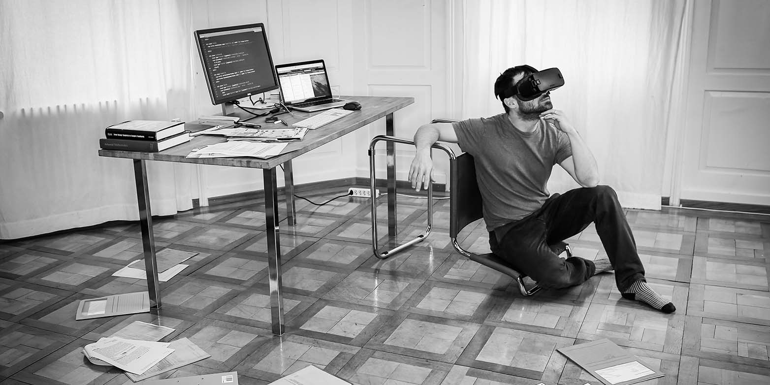Ein Mann mit VR Brille sitzt in einem Raum auf dem Fussboden, neben ihm steht ein Schreibtisch mit Computer