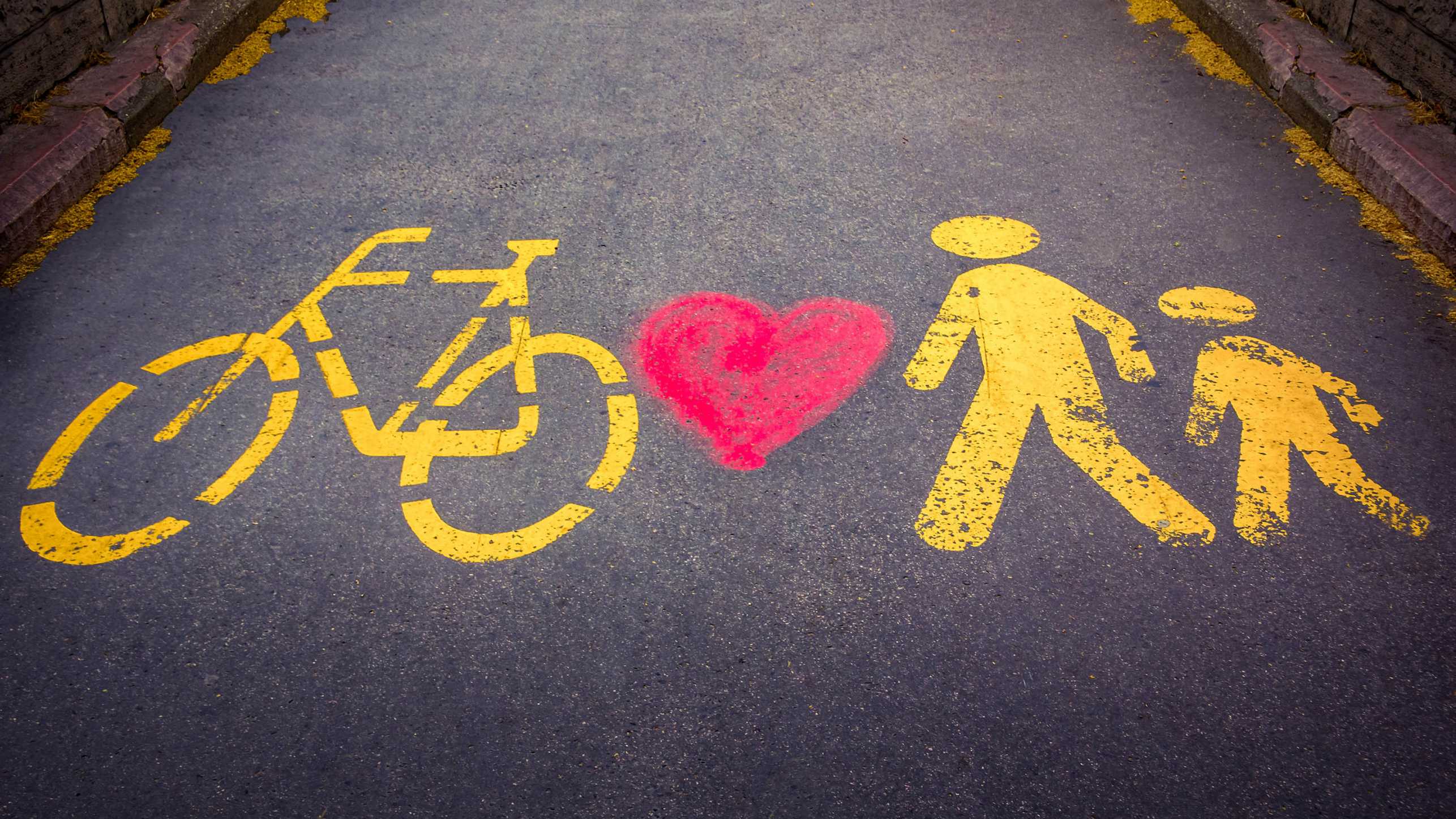 Auf einem gepflasterten Weg sind die Piktogramme von einem Fahrrad und zwei Personen in gelber Farbe und einem roten Herz zwischen Fahrrad und Personen gesprayt.