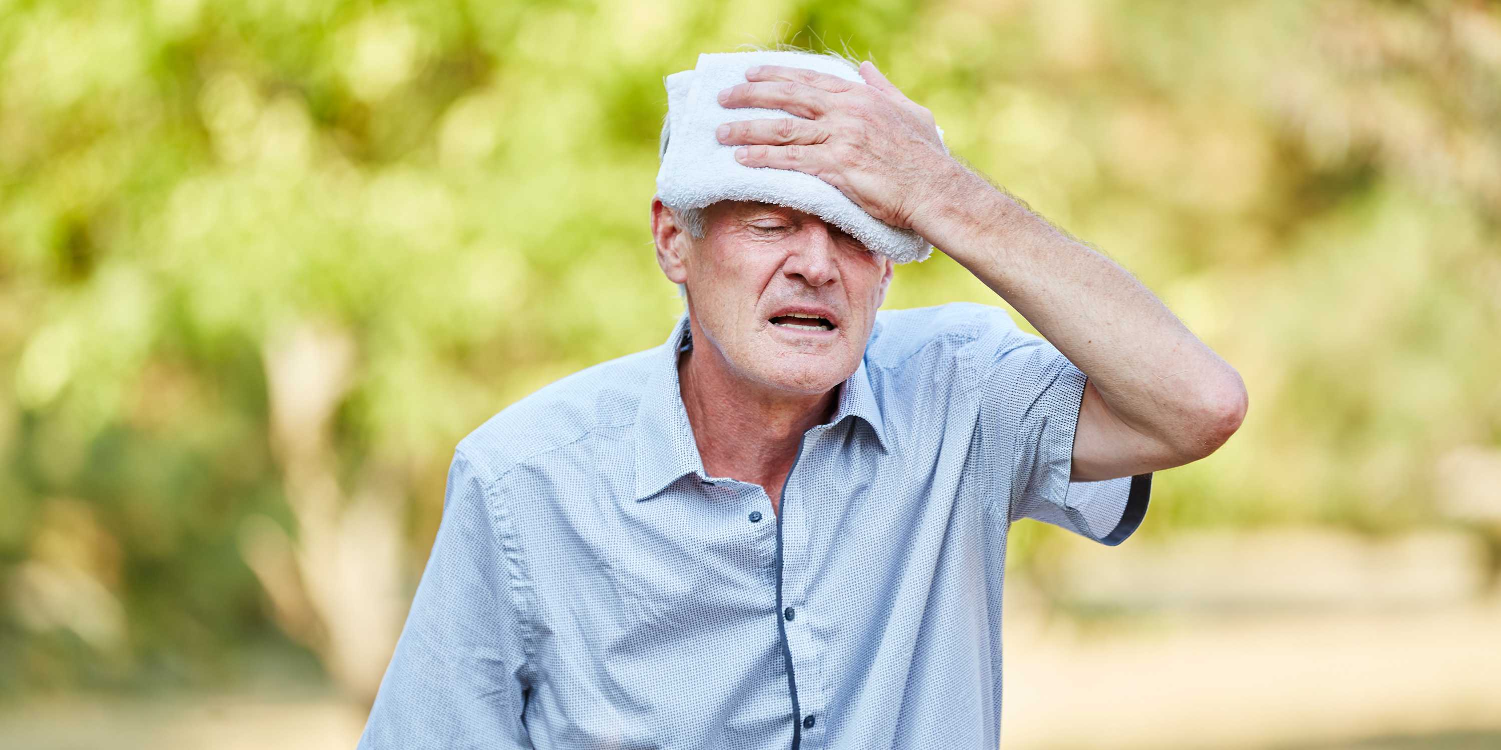 Ein alter Man hält sich ein feuchtes Tuch auf die Stirn und verzieht leidend das Gesicht.