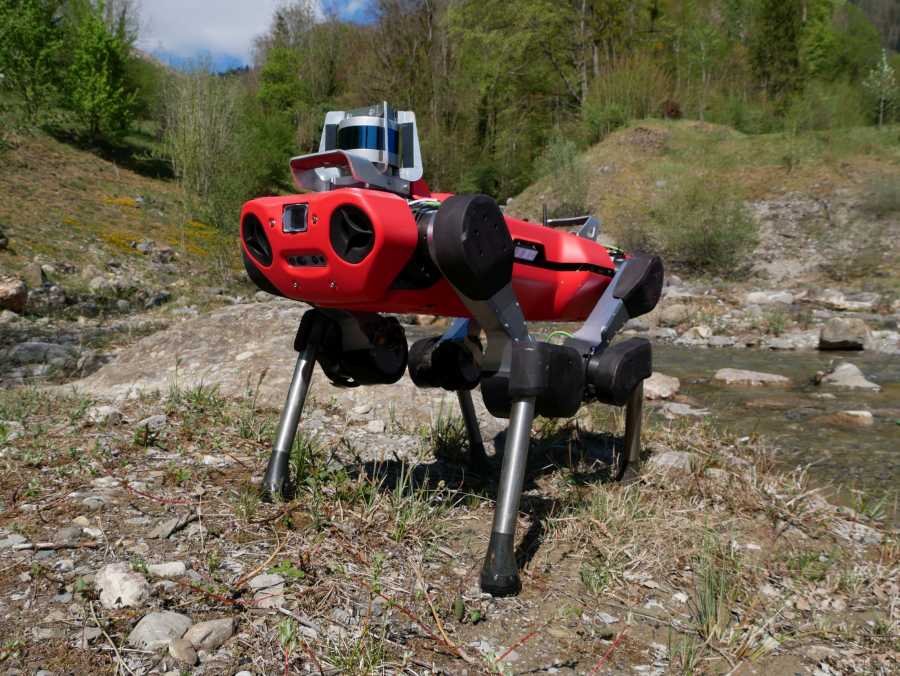 Vergr?sserte Ansicht: Roter vierbeiniger Roboter ANYmal in einer steinigen Umgebung, im Hintergrund Waldlandschaft.
