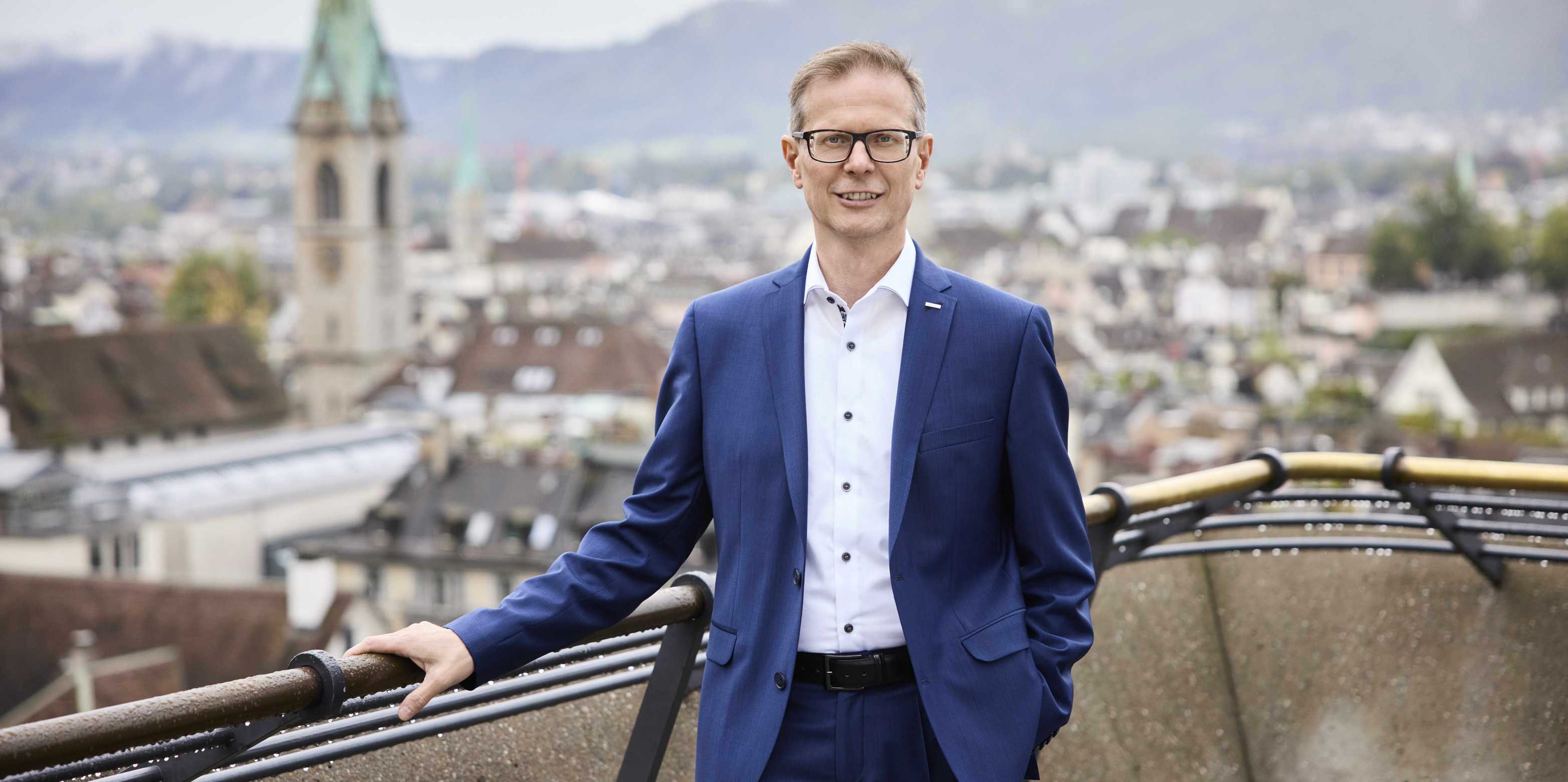Rektor Günther Dissertori trägt einen blauen Anzug und steht auf der Polyterasse. Im Hintergrund ist die Stadt Zürich zu sehen. 