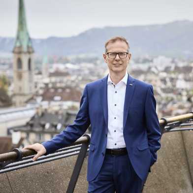 Rektor Günther Dissertori trägt einen blauen Anzug und steht auf der Polyterasse. Im Hintergrund ist die Stadt Zürich zu sehen. 